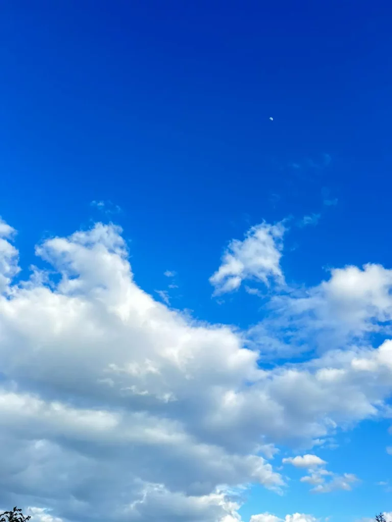 Un ciel bleu lumineux avec des nuages blancs épars au-dessus de collines verdoyantes et des arbres au premier plan. La lune est également visible en haut à droite, ajoutant une touche poétique à l'ensemble.