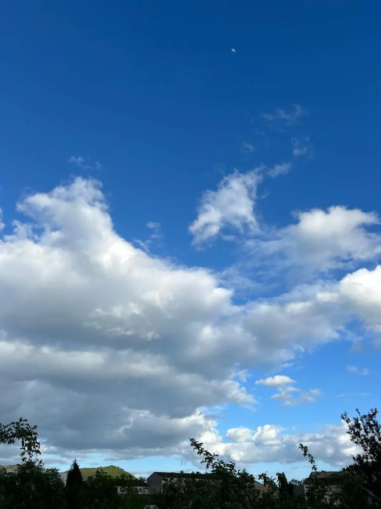 Un ciel bleu vif avec de gros nuages blancs et la lune visible en haut à droite. La scène est minimaliste, mettant en valeur la pureté et la douceur des nuages contre l'arrière-plan bleu profond.